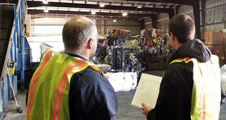 Two men in safety vests look on towards stacks of recycled material inside a recycling facility.