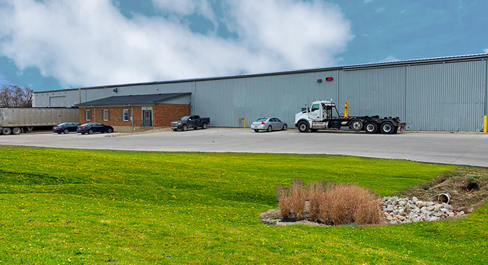 Large grey industrial building with multiple cars and tractor trailers parked outside.